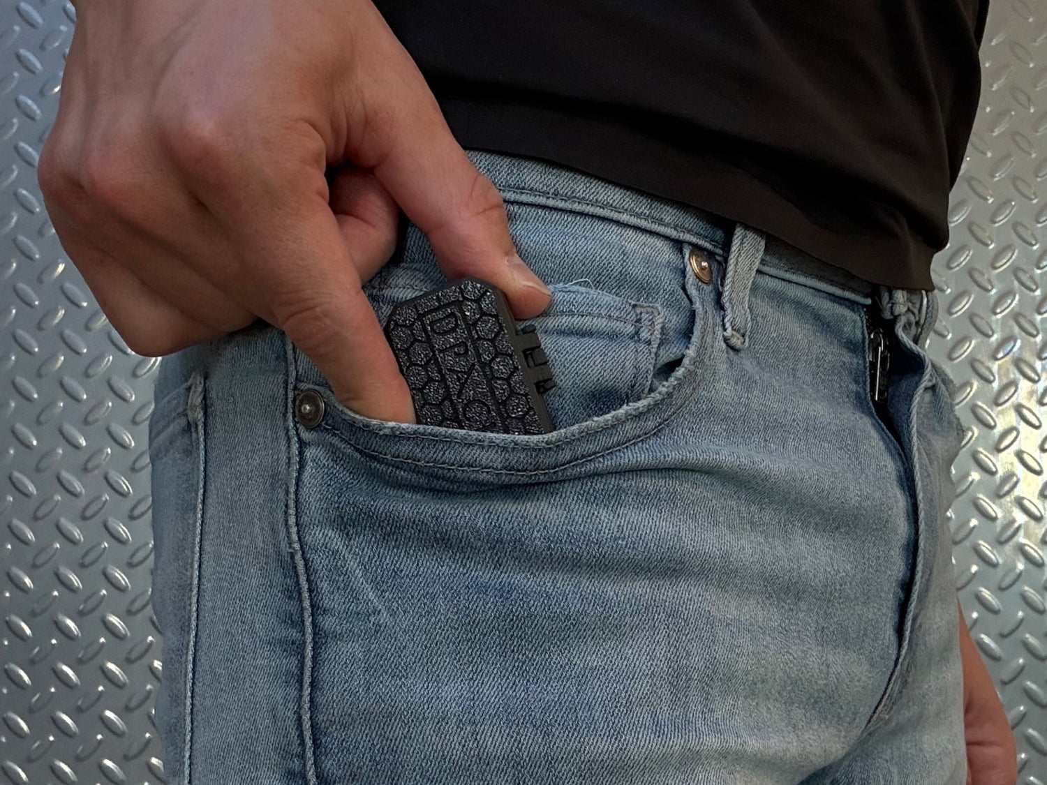 Person wearing light blue jeans with a black shirt, standing in a room with a metallic wall with Dpack in their pocket.